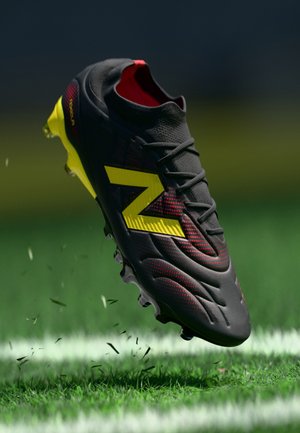 Zwart-rode voetbalschoen met gele accenten, textuurmateriaal, verhoogde patronen en noppen onder de zool, zwevend boven gras.