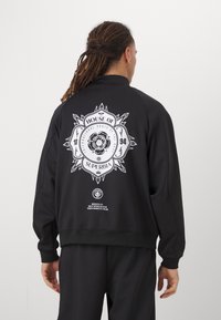 Svart hettegenser med et stort hvitt grafisk design på ryggen, som inneholder tekst og et løve-emblem. Laget av glatt stoff, avslappet passform.