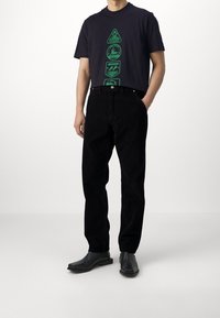 Černé kordové kalhoty s volným střihem, doplněné tmavě modrým tričkem s grafickými logy v zelené barvě. Outfit je doplněn černými botami.
