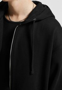 Svart zip-up hoodie med dragsko på huvan och silverfärgad dragkedja, med en mjuk textur och något lös passform. Bärs över en svart topp.