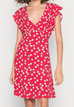 Mujer con vestido rojo con estampado de margaritas blancas y mangas cortas con volantes, de pie contra un fondo liso y claro.