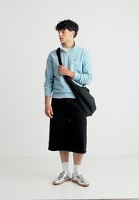 Νεαρός άνδρας που φοράει ανοιχτό μπλε πουλόβερ, μαύρες πλατιές βερμούδες, ασημένια αθλητικά παπούτσια και κρατάει μία μαύρη τσάντα χιαστή, στέκεται σε λευκό φόντο.