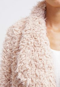 Texturerad, ljusrosa faux fur-sjal med en fluffig, inbjudande yta och öppen framdesign, draperad över en vit topp.