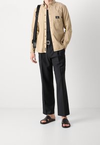 Béžová košile na knoflíky, černé kalhoty a černé sandály. Košile má kapsu na prsou; kalhoty jsou s rovnými nohavicemi. Minimalistický design.