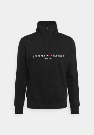 Hanorac negru cu fermoar dintr-un material moale, având guler înalt, buzunar frontal și logo-ul brodat "TOMMY HILFIGER EST. 1985".