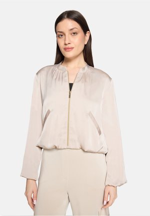 Kvinde iført beige satin bomberjakke med lynlås foran og matchende beige bukser, stående mod en ensfarvet lys baggrund.