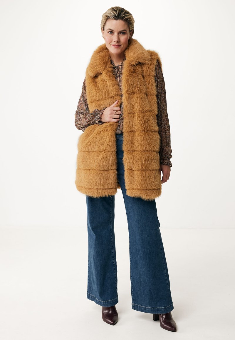 Chaleco de piel sintética en un tono marrón cálido, con un collar alto y textura a rayas. Combinado con jeans de pierna ancha y zapatos de tacón oscuros.