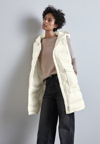 Béžová péřová vesta s otevřeným předním dílem, má kapuci a boční kapsy. Pod ní jsou vidět béžový svetr a tmavé džíny s vysokým pasem.