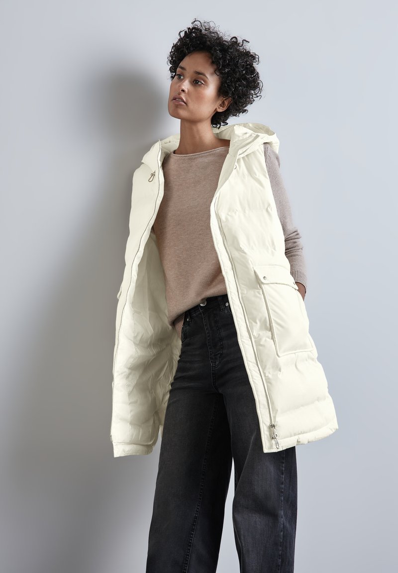 Béžová péřová vesta s otevřeným předním dílem, má kapuci a boční kapsy. Pod ní jsou vidět béžový svetr a tmavé džíny s vysokým pasem.