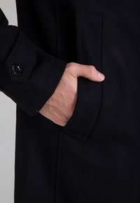 Cappotto di lana nera con polsini abbottonati e tasca laterale, caratterizzato da una trama liscia e una vestibilità su misura. Mano inserita nella tasca.