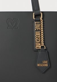 Mustast nahkatoos, millel on reljeefne südamekujuline logo ja kuldsed "Love Moschino" sildid, mis ripuvad käepidemest.