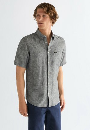 Homme portant une chemise grise à manches courtes avec une poche poitrine et un pantalon bleu marine, debout devant un fond blanc uni.