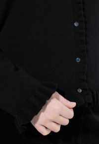 Černý cardigan s nařasenými okraji a stříbrnými patentkami. Měkká texturovaná látka viditelná blízko ruky, která upravuje zapínání.