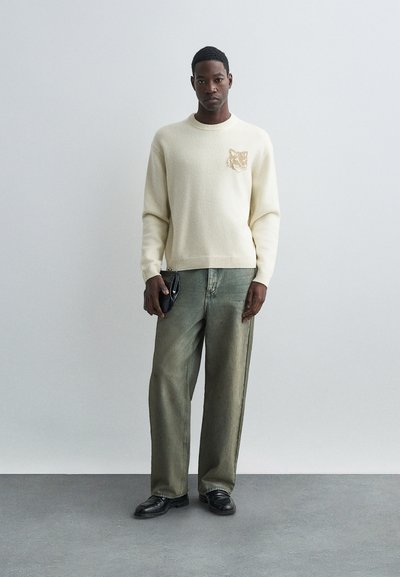 Fiatal férfi, aki egy krémszínű pulóvert visel, amelyen farkas arca van, laza zöldes farmernadrágot és fekete cipőt hord, és egy fekete textúrájú clutch-ot tart.