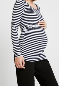 Randig långärmad topp i marinblått och vitt, med en figurnära design som rymmer en gravid silhuett, i kombination med svarta byxor.