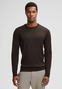 Dunkelbrauner Strickpullover mit langen Ärmeln, geripptem Rundhalsauschnitt und Bündchen. Glatte Oberfläche, taillierte Passform, kombiniert mit hellgrauen Hosen.
