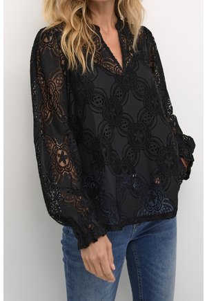 Kvinde iført en sort blondetop med lange ærmer og blomsterprint, kombineret med blå jeans, stående mod en ensfarvet hvid baggrund.