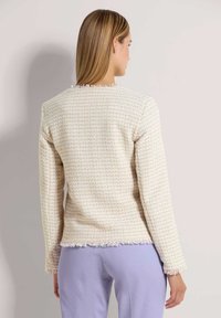 Crèmekleurige tweedjas met kleurrijke spikkels, structuurweefsel, lange mouwen en franjes aan de randen, gedragen over lavendelkleurige broek.