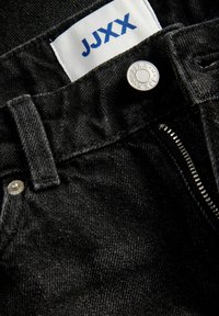 Μαύρο τζιν από denim με μερικώς ανοιχτό φερμουάρ, ασημένιο κουμπί με χαραγμένα τα γράμματα JJJX και λευκή υφασμάτινη ετικέτα με μπλε κείμενο JJJX.