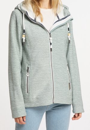 Kvinde iført en lysegrå hættetrøje med lynlås og hvide snore samt to lynlåslommer foran over en hvid skjorte og lyseblå jeans.