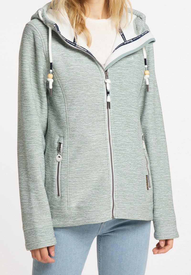 Femme portant un sweat à capuche gris clair avec fermeture éclair, cordons blancs et deux poches frontales zippées, sur un t-shirt blanc et un jean bleu clair.