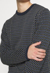 Marinblå stickad tröja med en texturerad design som har små vita och gula hjärtformer. Ribbat nacke och muddar, medelhård vikt.