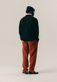 Man in een zwarte baret, zwarte sweater, roestkleurige broek en zwarte schoenen, staand met de rug naar de camera tegen een eenvoudige beige achtergrond.