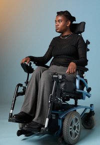 Cadeira de rodas elétrica com assento em tecido cinza escuro, apoios de braços ajustáveis e pneus traseiros robustos. O utilizador veste uma blusa preta com riscas e calças cinzas.