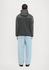 Sort hætte sweatshirt med geometrisk mønster på skuldrene, grå ryg, parret med løse lyseblå jeans og sorte sko. Ensfarvet hvid baggrund.