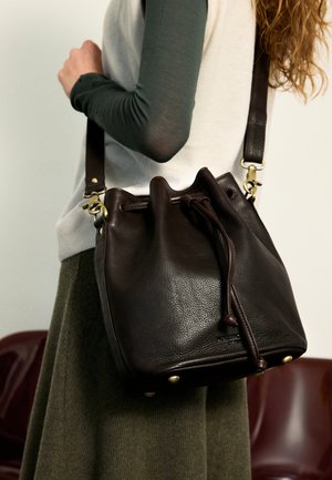 Person trägt grünes langärmeliges Shirt und grünen Rock und hält eine dunkelbraune Leder-Bucket-Tasche mit Kordelzug und Schulterriemen.