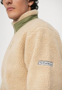 Beige fleece pullover met een groene kraag, voorzien van een rits en een "No Problemo" label op de linkermouw. Getextureerd, zacht materiaal.