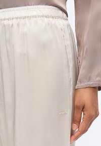 Ελαστικό παντελόνι κρεμ χρώματος με μαλακή μέση, συνδυασμένο με μακρυμάνικη τοπ σε σκούρο γκρι-καφέ απόχρωση, χέρι που ακουμπάει δίπλα στο ισχίο και μικρό λογότυπο "BOSS" ορατό.