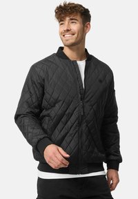 Veste bomber matelassée noire avec fermeture éclair, poignets et col côtelés. Elle présente une texture lisse et un petit logo sur la poitrine.