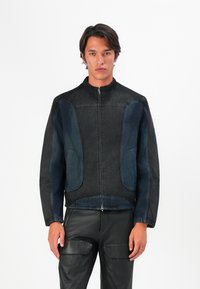 Jauns vīrietis valkā melnu un tumši zilu rāvējslēdzēja jaku ar augstu apkakli un melnu ādas bikses, stāvot pretī baltai fona.