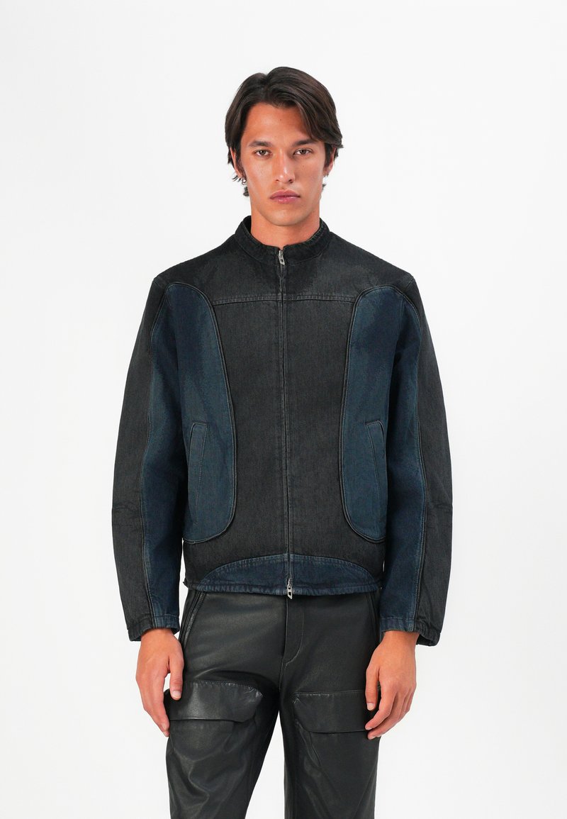 Jauns vīrietis valkā melnu un tumši zilu rāvējslēdzēja jaku ar augstu apkakli un melnu ādas bikses, stāvot pretī baltai fona.