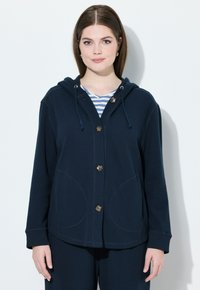 Giacca con cappuccio blu navy in morbido tessuto, con grandi tasche, apertura con bottoni e cappuccio regolabile; indossata sopra una maglietta a righe.