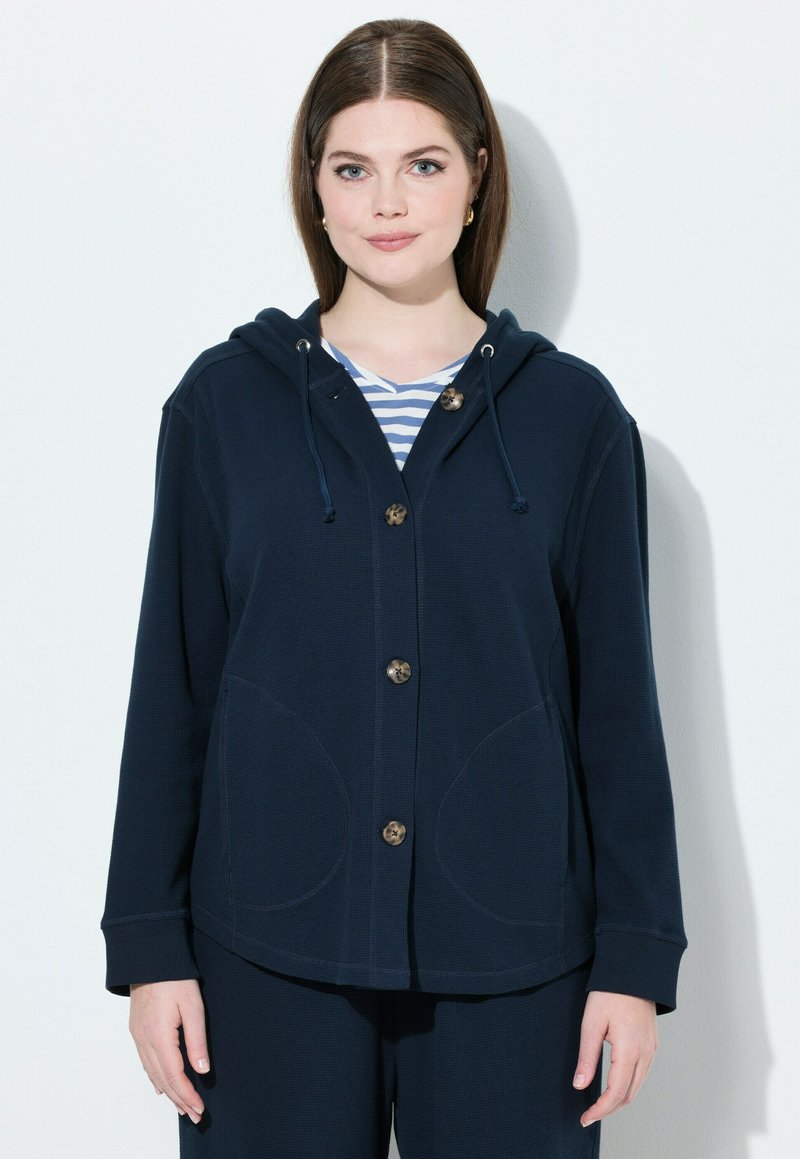 Giacca con cappuccio blu navy in morbido tessuto, con grandi tasche, apertura con bottoni e cappuccio regolabile; indossata sopra una maglietta a righe.