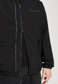 Svart, lätt bomberjacka med sidodragkedjor, elastiska ärmslut och en diskret blå logotypdesign på bröstet och sidopanelen.