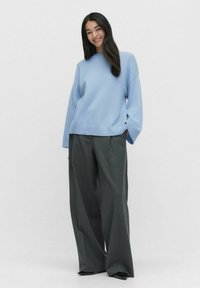 Jasnoniebieski, oversized sweter z prążkowanym dołem, w zestawieniu z luźnymi, szarymi spodniami w kratę i czarnymi butami. Gładka dzianinowa tekstura.