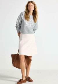 Grå sweatshirt med teksten "WEST COAST", cremet nederdel, brun tote bag og brune snørebåndssko. Afslappet outfit med løs pasform og forskellige teksturer.
