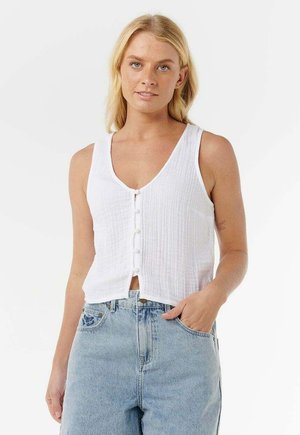 Młoda kobieta stojąca, ubrana w białą, bezrękawnikową, zapinaną na guziki bluzkę oraz jasnoniebieskie, wysokowciskane dżinsy, z jedną ręką w kieszeni.