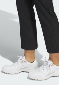 Λευκά αθλητικά παπούτσια με υφασμένες σόλες, ανοιχτογκρι λεπτομέρειες και λευκά κορδόνια, φορεμένα με μαύρο παντελόνι cropped, που αναδεικνύουν μια στενή εφαρμογή.