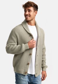 Cardigan de punto en gris claro, con un cuello chal, grandes botones, paneles texturizados y puños acanalados. Lleva encima una camisa blanca.