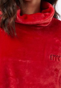 Felpa rossa in peluche con collo alto a cuffia, texture morbida e dettaglio del logo ricamato.