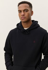 Μαύρη φούτερ με κουκούλα και κορδόνι, με μικρό κόκκινο λογότυπο «II» στο στήθος. Μαλακό ύφασμα, τσέπη καγκουρό και ριμπ μανσέτες.