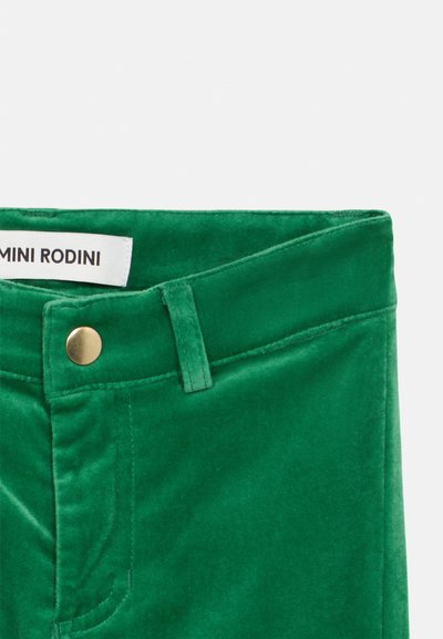 Pantaloni din catifea verde, având o textură netedă, talie îngustă și un detaliu cu un buton în ton de aur. Eticheta brandului "Mini Rodini" este vizibilă.