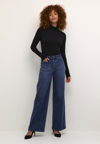 Traperice visokog struka širokih nogavica u tamnoj boji, s dva prednja džepa i zatvaranjem na gumbe. Kombinirane s crnim topom dugih rukava.