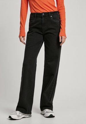 Jeans de mezclilla negra de pierna ancha con bolsillos frontales, trabillas para cinturón y cierre de botón metálico, combinados con una camiseta de manga larga naranja y zapatillas grises.