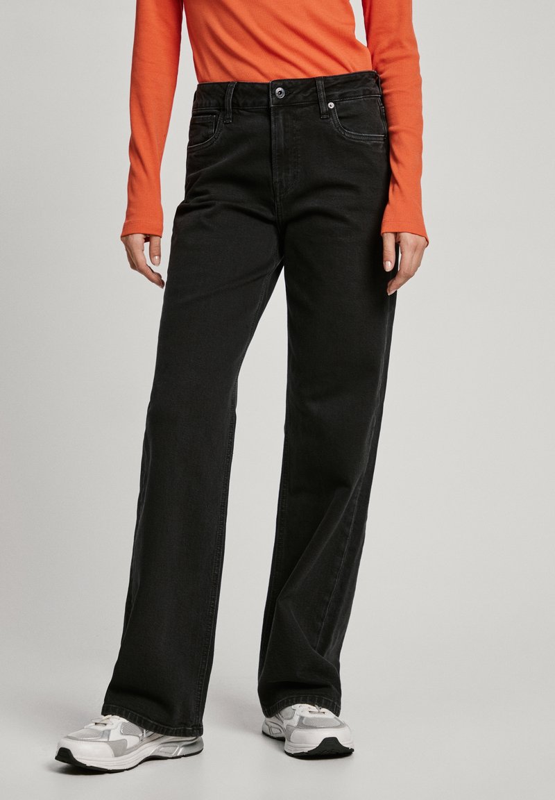 Svarta denim jeans med vida ben, framfickor, bälteshällor och metalldragknapp, tillsammans med en orange långärmad skjorta och grå sneakers.