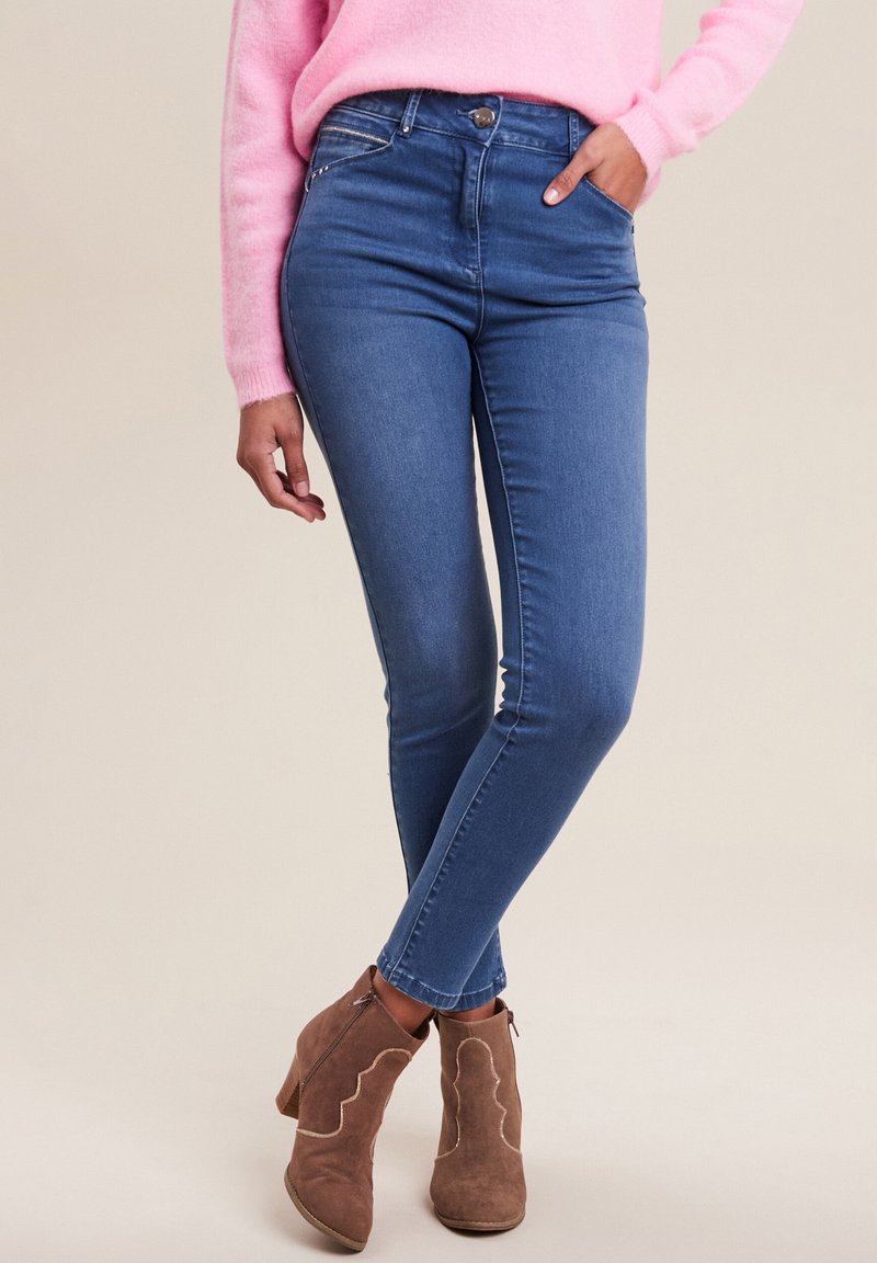 Jean bleu taille haute à coupe slim, en деним. Associés à des bottines brunes à motif ondulé et fermeture éclair sur le côté.